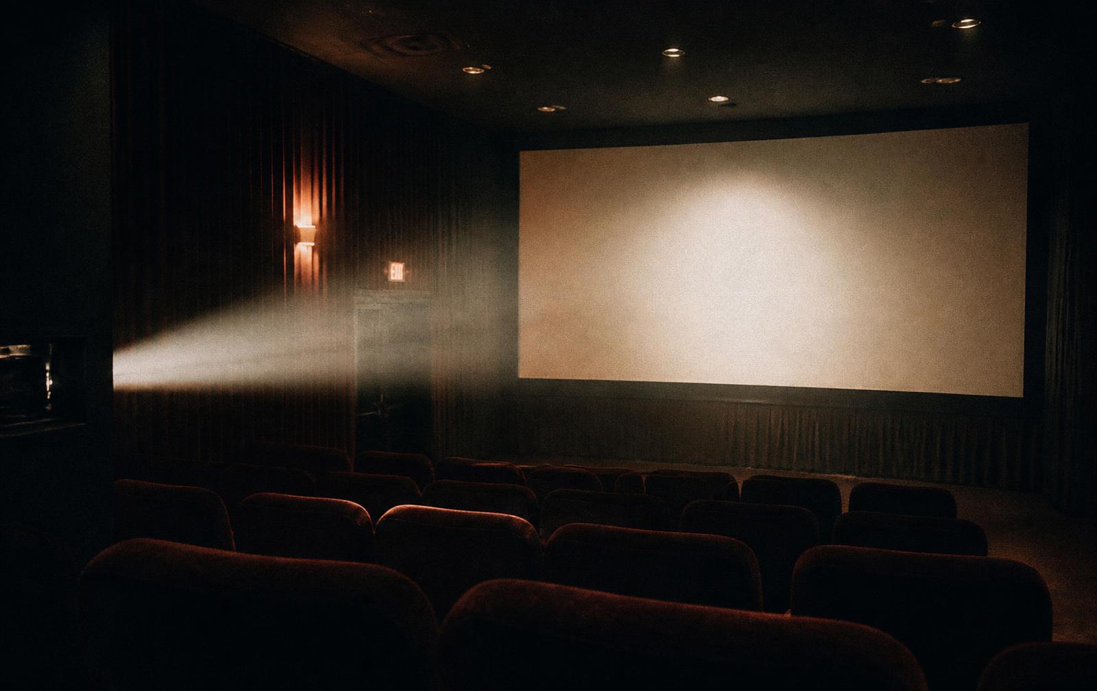An empty screening room with a projector beam crossing the haze toward a blank screen.